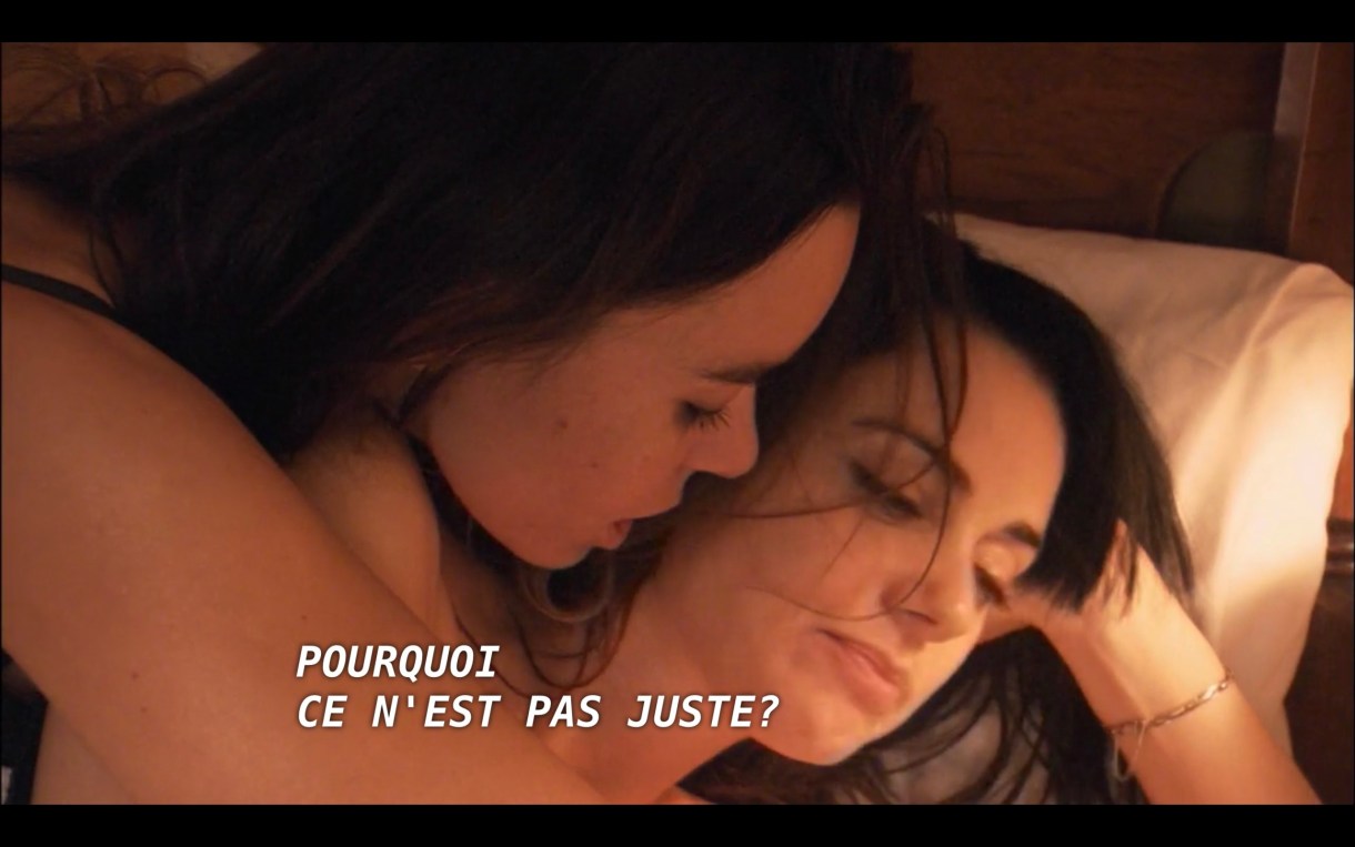 Jenny lays on her side in a bed, her hand propping up her head. A brunette woman (who Jenny just met) leans over her, saying in French, "Pourquoi ce n'est pas juste?"