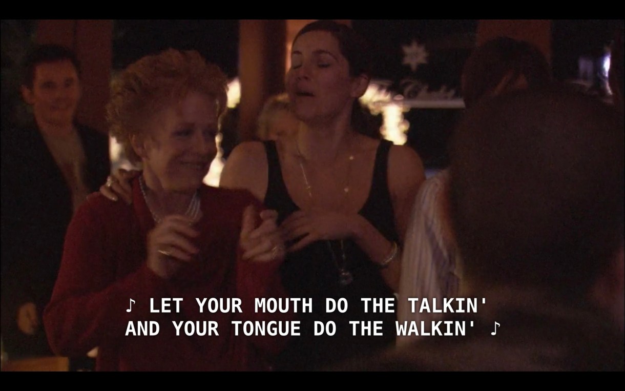 Peggy (wearing a red sweater and a pearl necklace) and Helena (wearing a black dress and long gold necklace) stand next to each other as they watch the raunchy song performance. The singers, off screen, sing, "Let your mouth do the talkin' and your tongue do the walkin'"