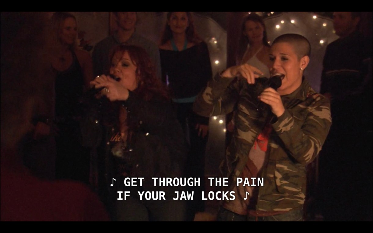 Famed singers, God-des (who has a shaved head and is wearing a camouflage jacket) and She (who has curly red hair and wearing a black jacket), sing, "Get through the pain if your jaw locks."