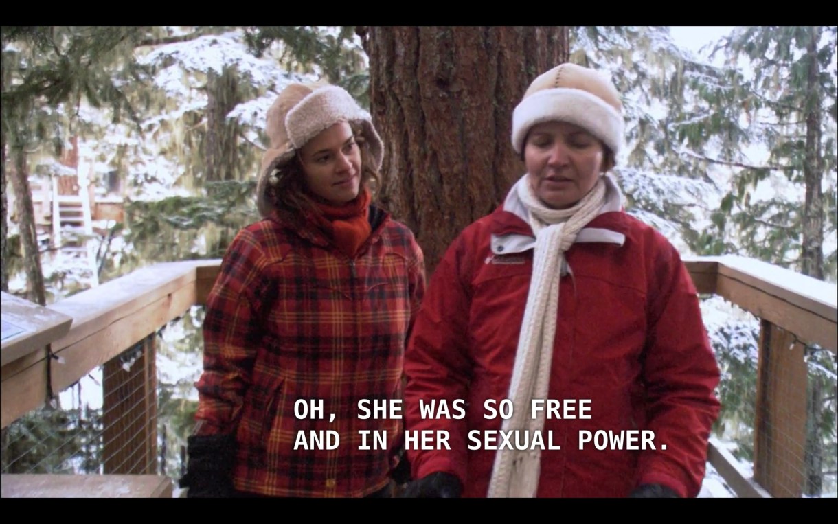 Alice (wearing a red plaid winter jacket and a brown ear-flap hat) walks next to Marilyn (a woman Alice just met, wearing a red winter jacket and white scarf) on a wooden deck in the forest of snow-covered evergreen trees. Marilyn says, "Oh, she was so free and in her sexual power."