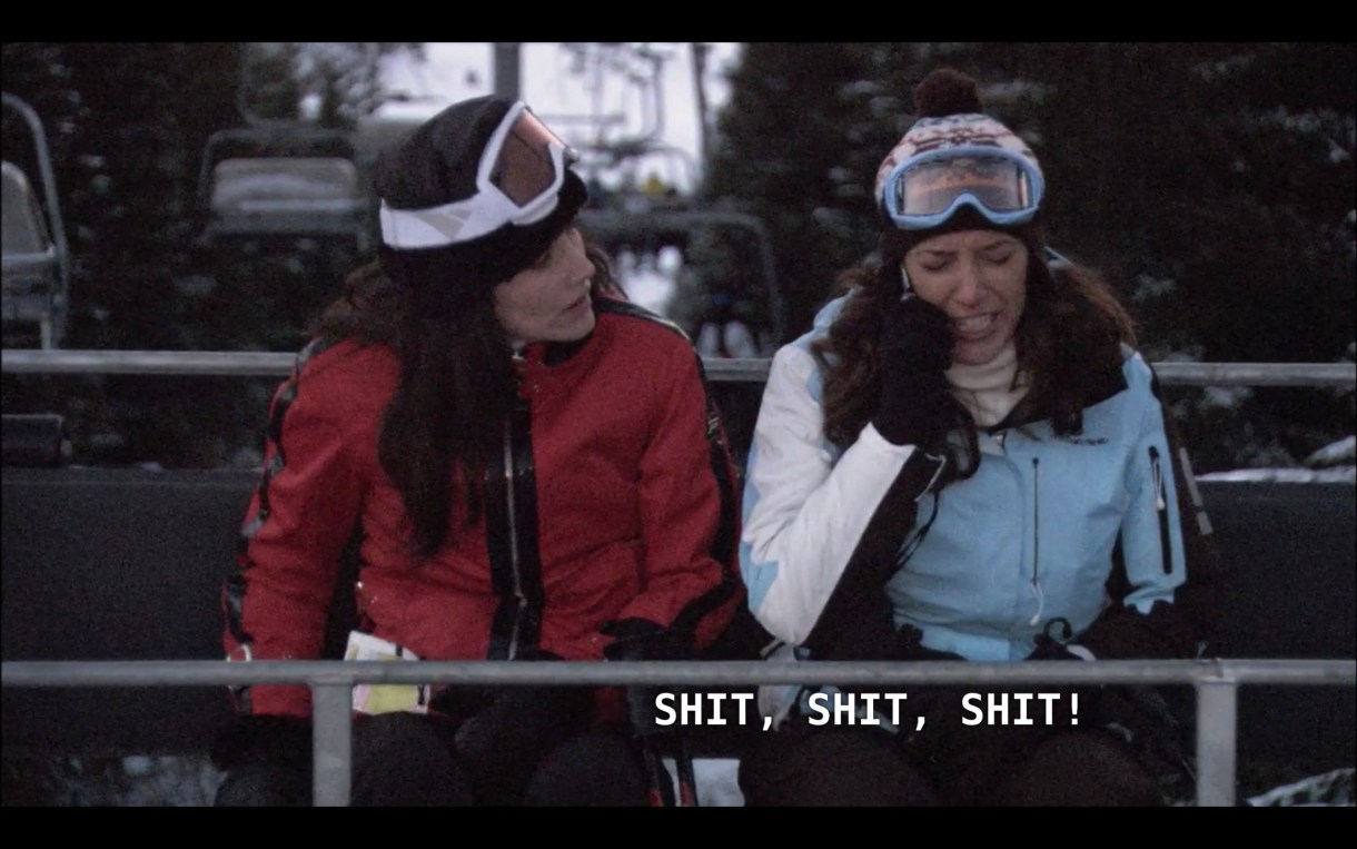 Helena (wearing a red ski jacket, black hat, and ski goggles) and Bette (wearing a blue and white ski jacket, hat, and goggles) on a chairlift. Bette is listening to her voicemail and says, "Shit, shit, shit!"