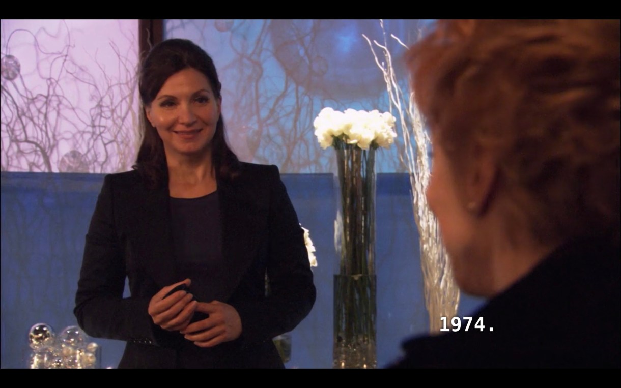 Marilyn (wearing a black blazer, her brown hair tied back) stands in front of a window and a vase of very tall white roses. She is looking incredulously at Peggy, whose back is to the camera. Peggy says to her, "1974."