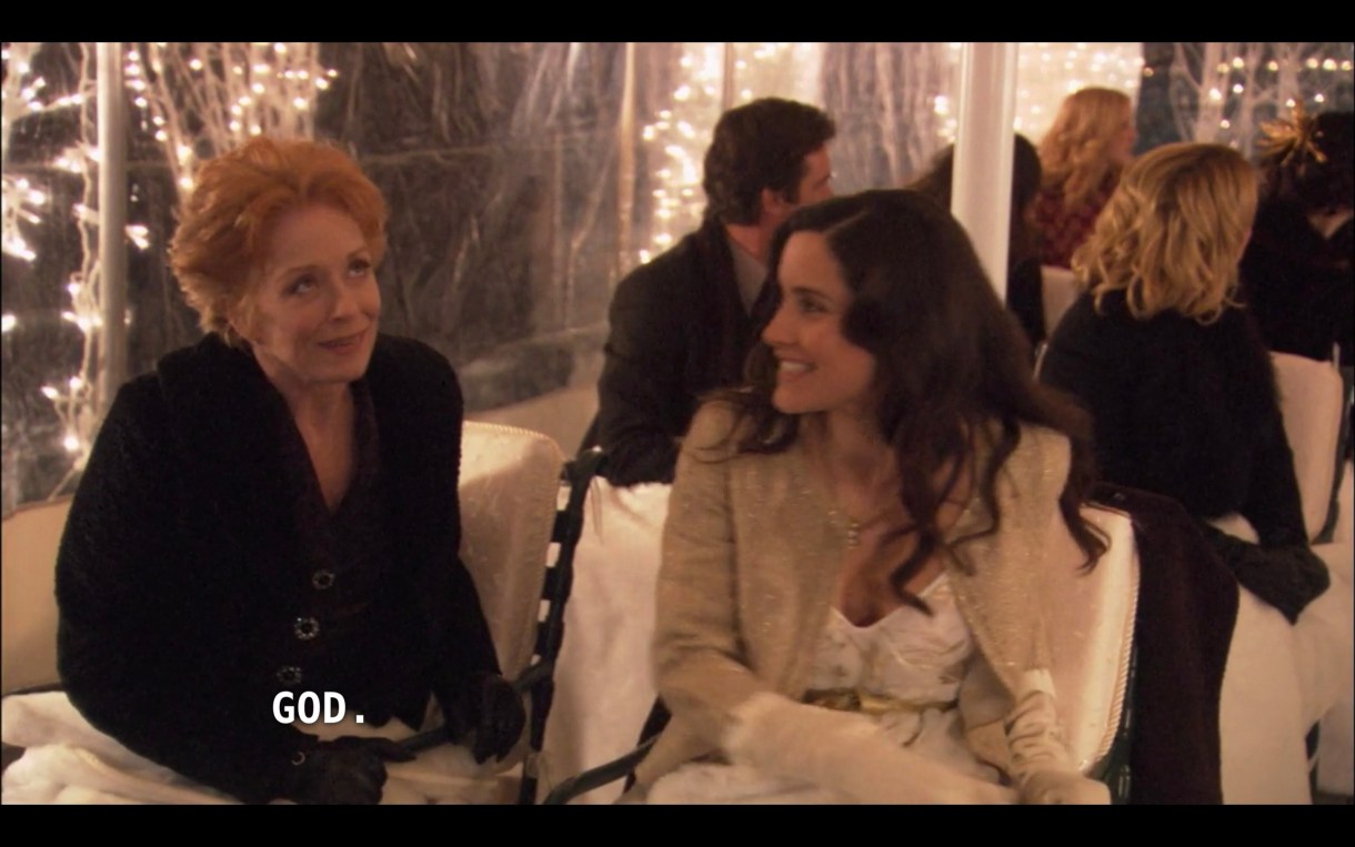 Peggy (wearing a black jacket) and Helena (wearing a white top under a tan jacket) sit in tent for Carmen and Shane's wedding. Tina and Henry, and other guest, are sitting behind them. Peggy says, "God."