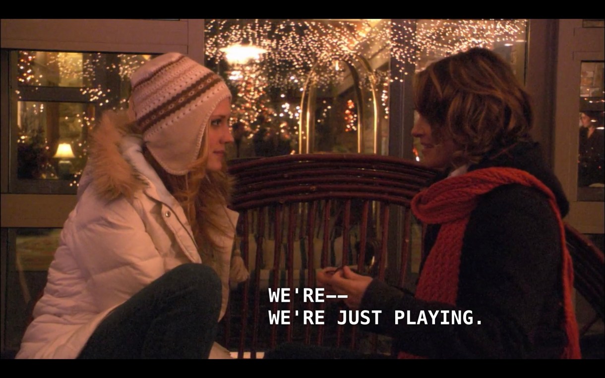 Lara (wearing a white puffy coat and a tan hat) and Alice (wearing a black jacket and red scarf) sit on a bench in front of a big room with tons of twinkly lights hanging from the ceiling. Alice says, "We're -- we're just playing."