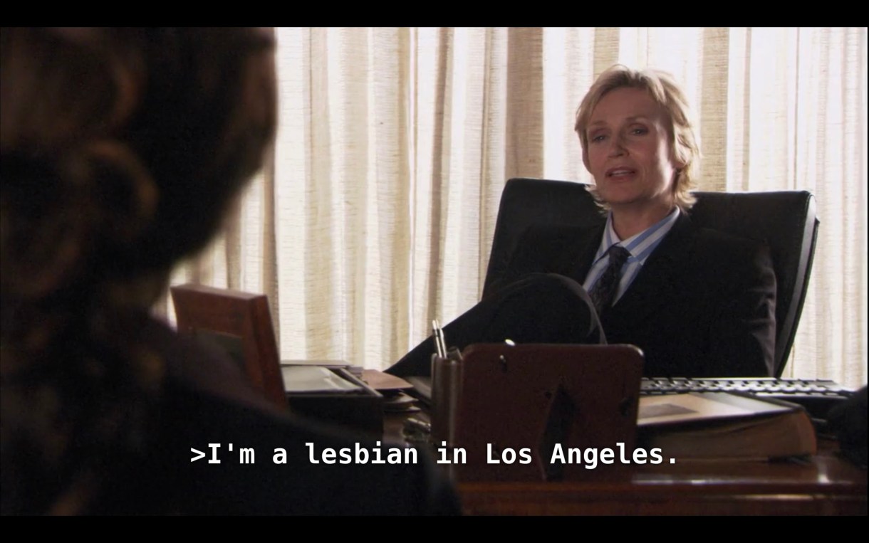 Joyce (the lesbian lawyer played by Jane Lynch) is sitting behind her desk and wearing a black blazer over a blue-and-white shirt and a tie. She says to Bette (who is sitting across from her, her back to the camera), "I'm a lesbian in Los Angeles."