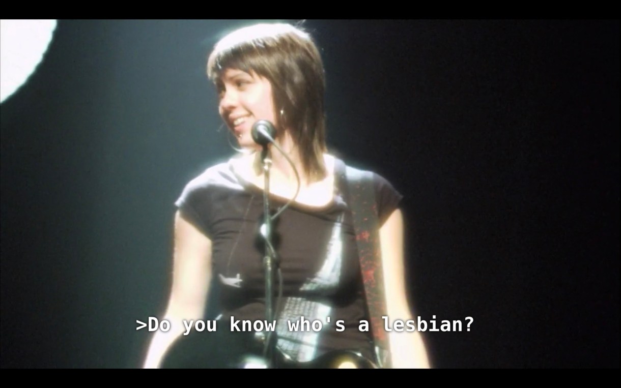 Tegan (or Sara? Hard to say) Quin is wearing a black shirt and standing at a microphone on stage. She asks, "Do you know who's a lesbian?"