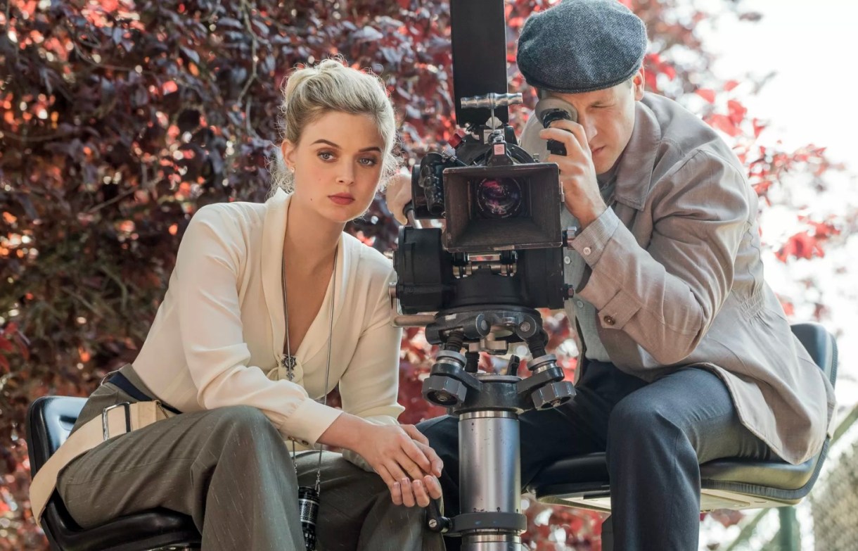 Image: A blonde white woman in a v-neck white silk shirt and grey pants is sitting behind a camera with a white man, who is looking through the camera. This scene appears to take place in the 40s or 50s.