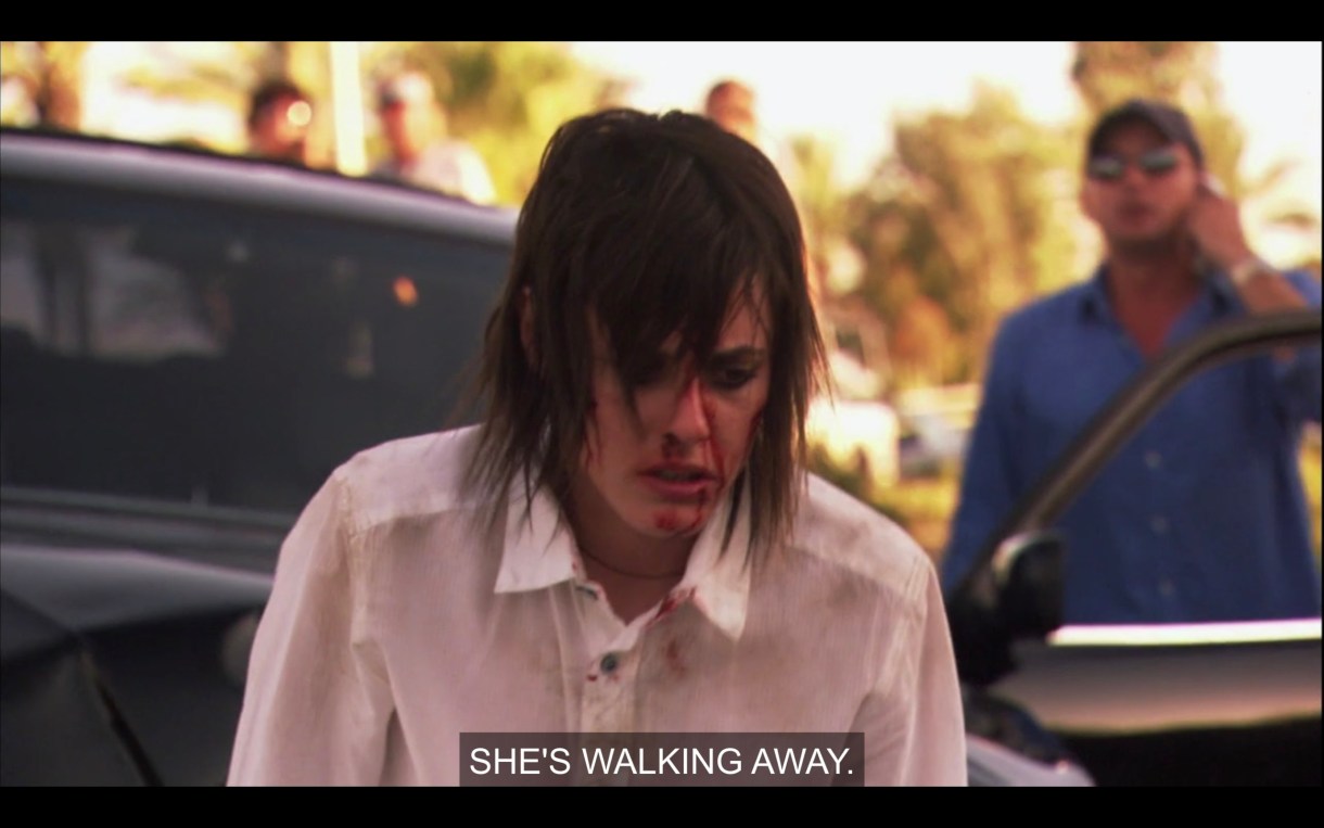 Shane, in a white button-up shirt with blood all over her face and her hair all over her face is walking away from a car crashed she caused. in the background a dude is on the phone saying "She's walking away."