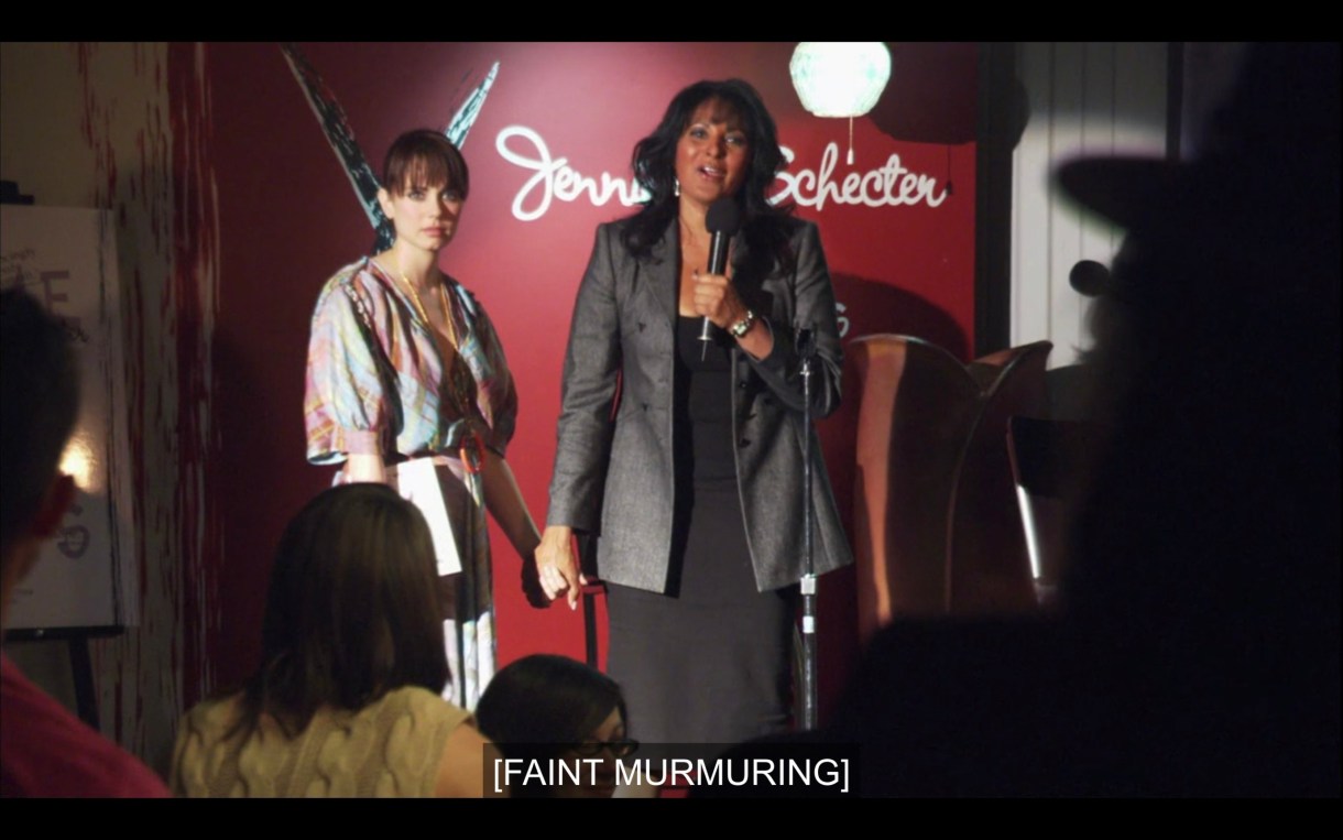 On the stage for her reading. Kit in an all-black ensemble is on the mic, Jenny in a flowing dress is next to her. Kit is holding Jenny's hands. Jenny looks like she just saw a ghost. Caption reads: [faint murmuring]