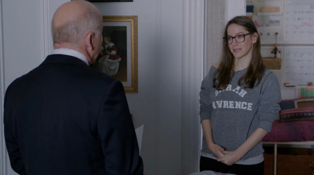 Image: the lesbian chief-of staff, a white woman in glasses with long dark hair, wearing a Sarah Lawrence sweatshirt, looks at her boss. His back is to us but he's clearly a bald white man.