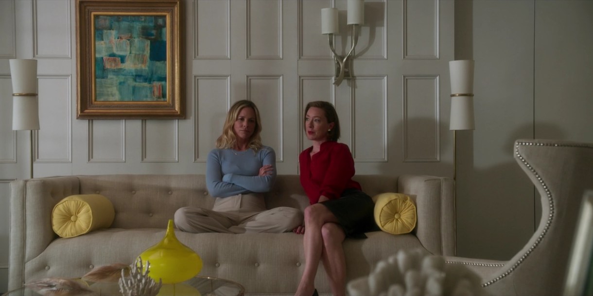 Image: A living room that exudes an air of wealth. White walls, off-whtie sofa, white lighting fixtures, some yellow accents, a gold-framed photo. On the sofa sit two white women. One is wearing a skirt her legs, bare from the knees, are crossed with her feet on the floor, and is leaning slightly towards the other, who is relaxed and sitting cross-legged on the couch in a blue sweater. She is looking at the camera.
