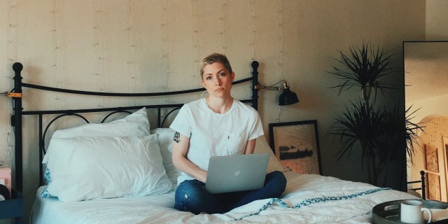 A woman sits cross-legged on her bed with a laptop. She is dead inside.