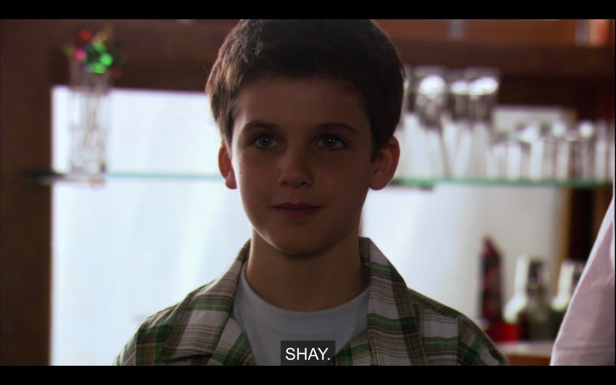 Shay stands shyly wearing a green and white plaid shirt. He introduces himself, saying, "Shay."
