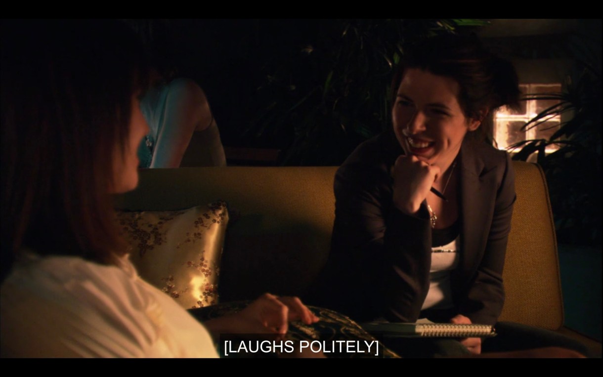 A journalist in a blazer chats with Jenny on a couch at her reading. Subtitles say, "[Laughs politely.]"