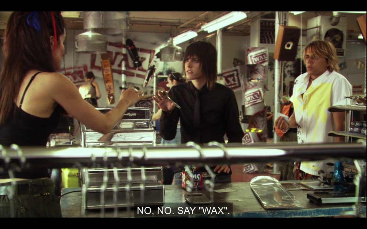 Helena is on her first day as receptionist at Wax. She's talking on the phone. Shane puts her hand out to Helena in a "stop" gesture and says, "No, no. Say 'Wax'"