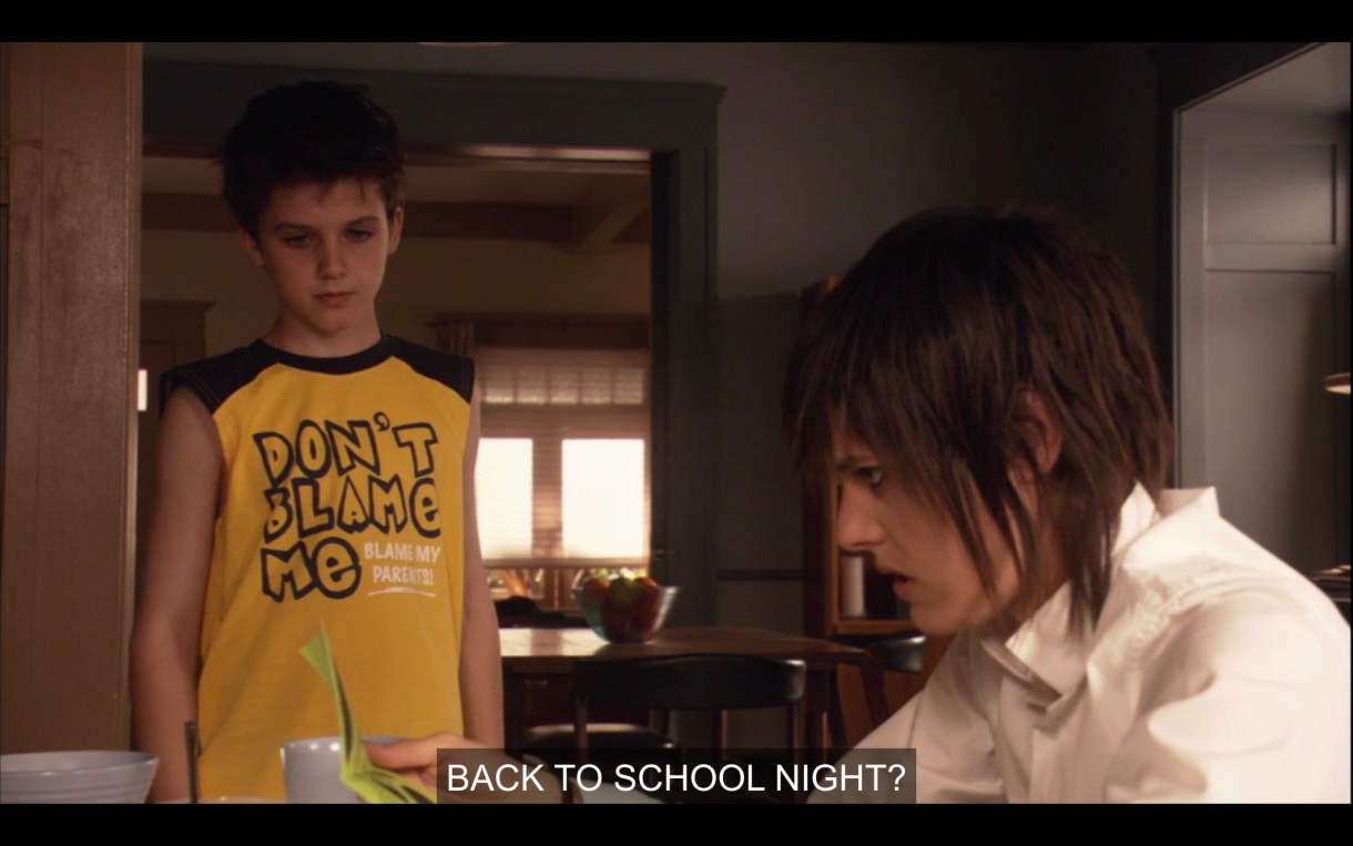 Shay (wearing a yellow tank top that reads, "Don't blame me, blame my parents!") has handed Shane a green flyer from school. Shane is sitting at the kitchen table. Shane looks at it and says, "Back to school night?"