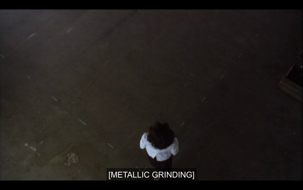 Overhead shot of Bette standing on a dark concrete floor as she enters Jodi's studio. Subtitles read, "[Metallic grinding]"