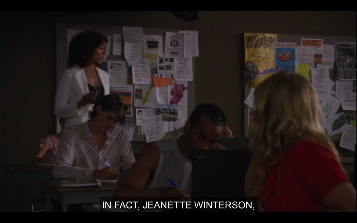 Bette is wearing a bright white blazer and pacing around the classroom giving a lecture. She says, "In fact, Jeanette Winterson..."