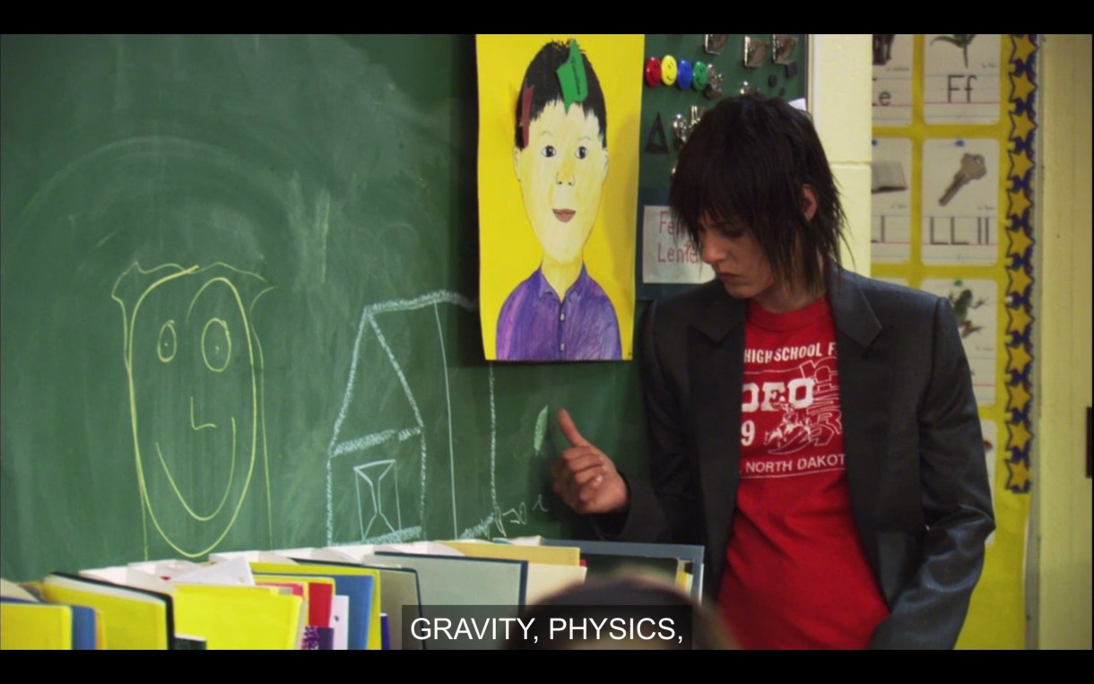 Shane (wearing a red t-shirt under a black blazer) leans against a green chalkboard in Shay's classroom. There is a smiley face and a house drawn on it. 