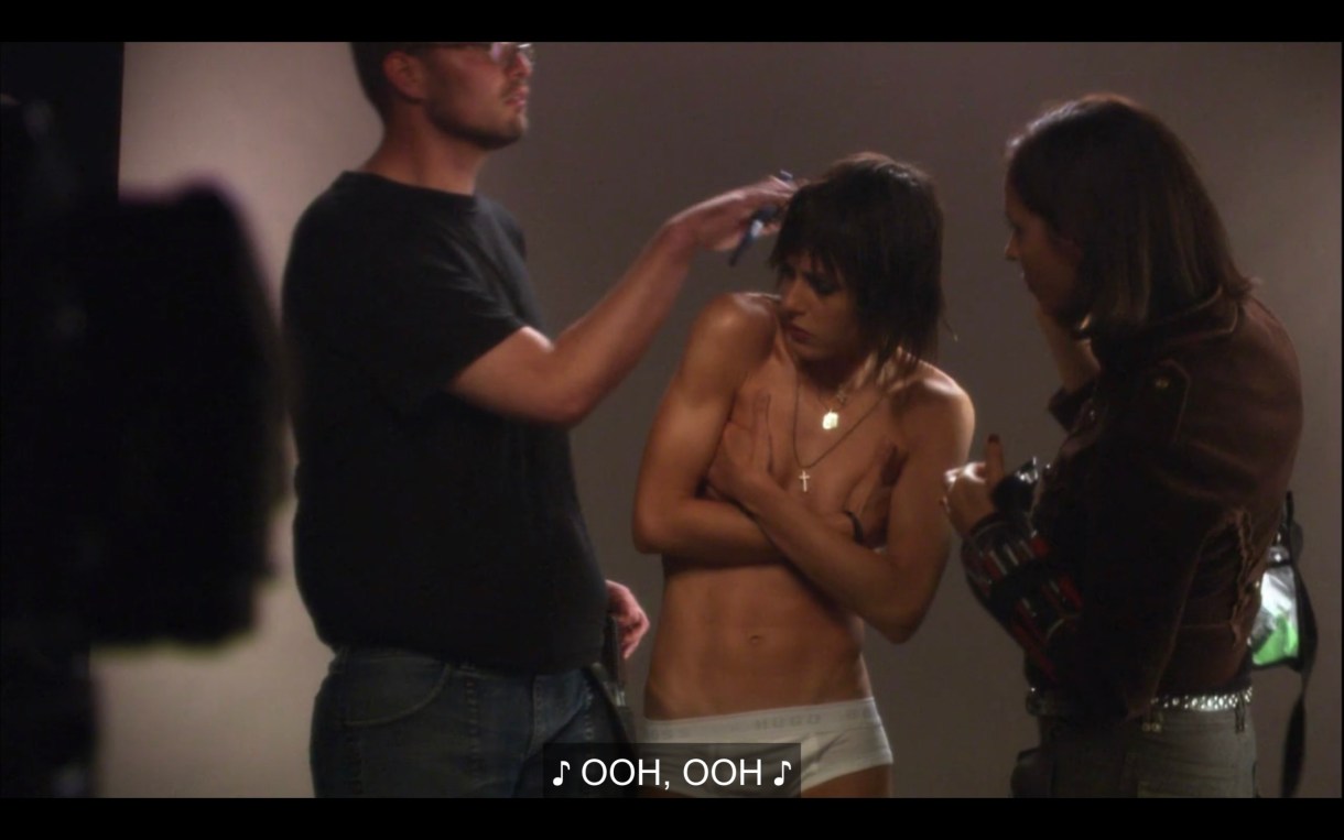 Shane stands in white brief underwear and topless for a photoshoot. Her hands are covering her boobs, as two people prep her for a photoshoot.