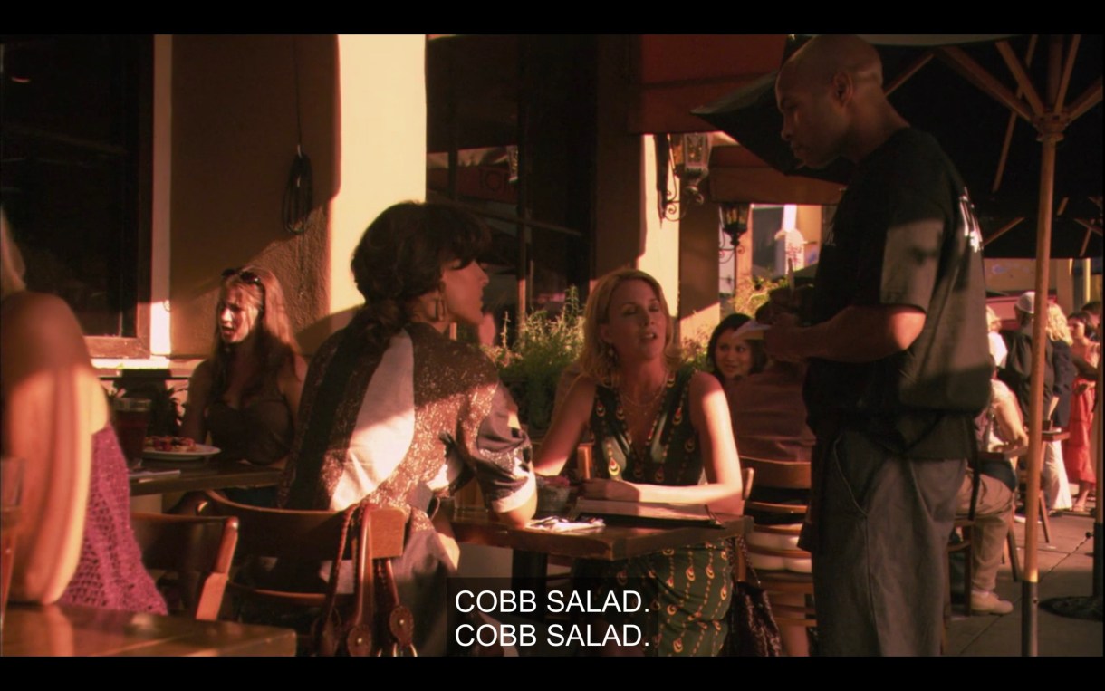 Tina and Bette sit at an outdoor restaurant table. They both order a Cobb salad from the waiter standing next to them.