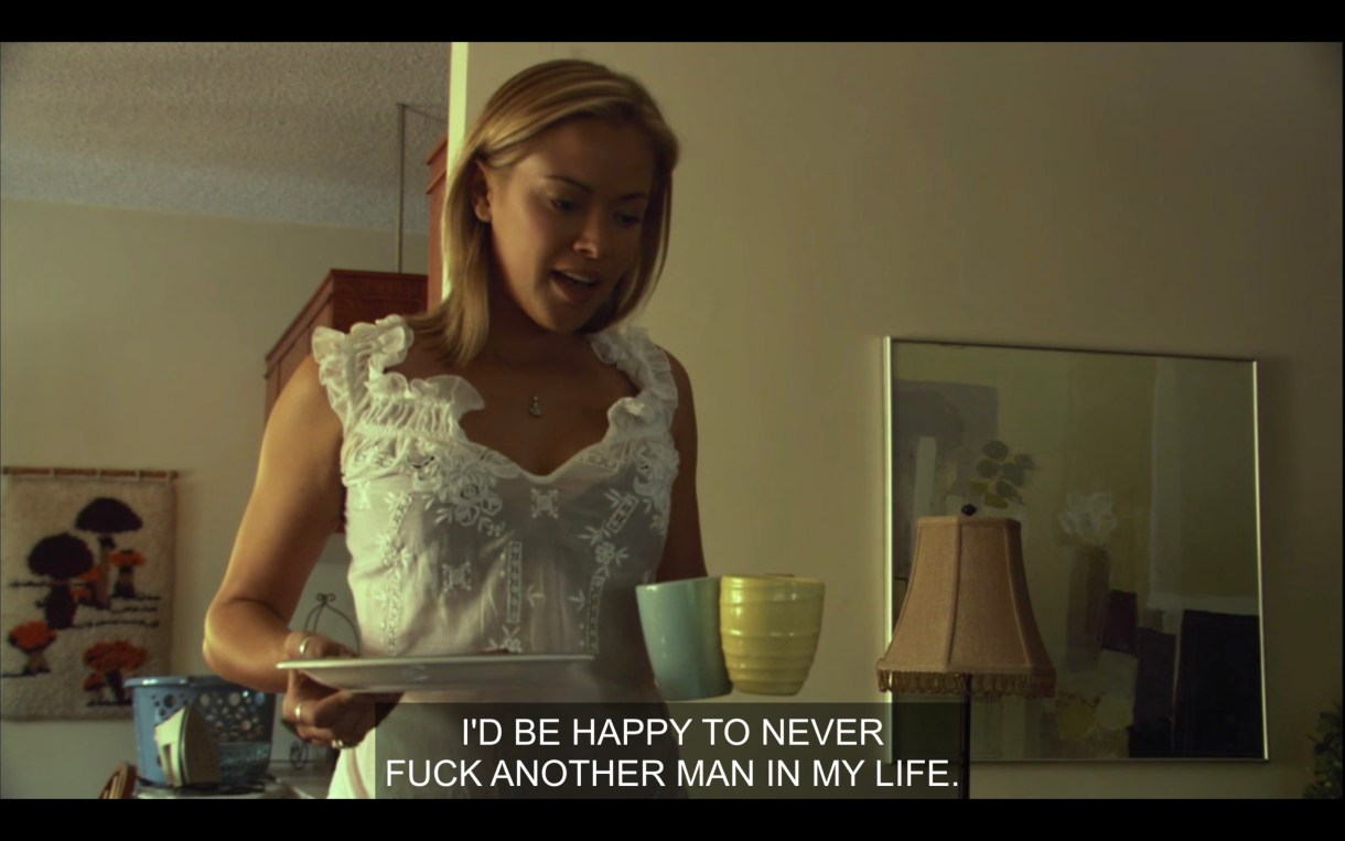 Paige (wearing a white blouse) holds a plate and two coffee mugs in her hands. She says, "I'd be happy to never fuck another man in my life." 