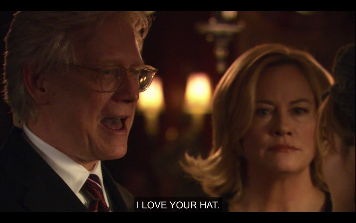 An old white guy with glasses and a suit stands next to Phyllis in a dimly lit room. "I love your hat."