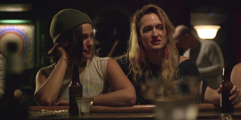 Two women sit at a bar with beer bottles.