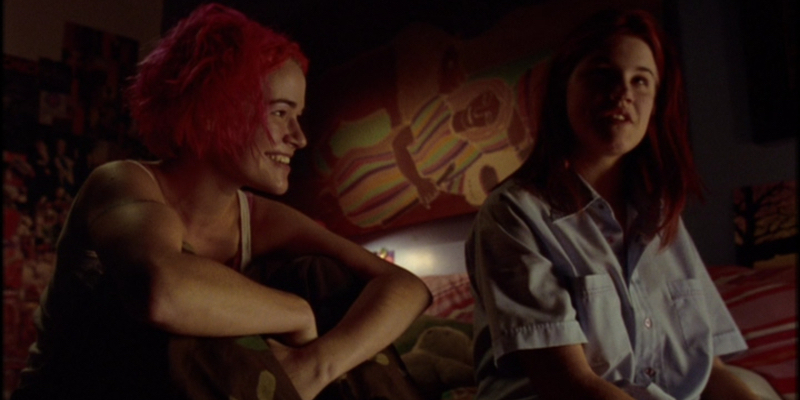 A girl with pink hair looks at another girl while they sit next to each other on a bed.