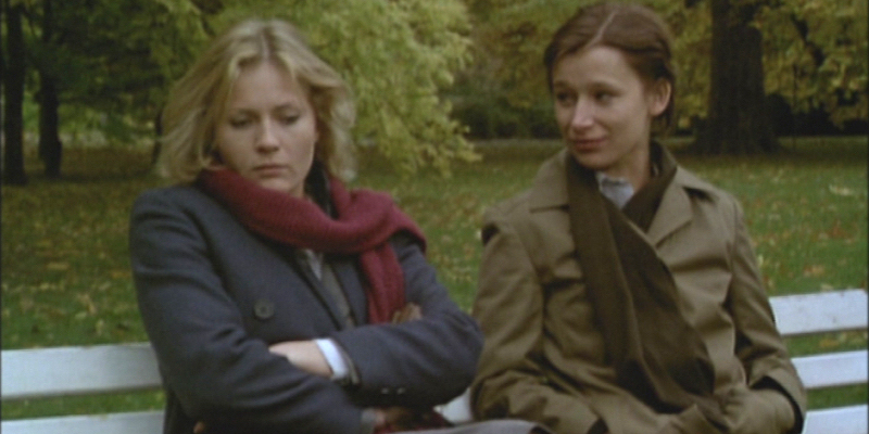 Two women wearing coats and scarves sit next to each other on a park bench.