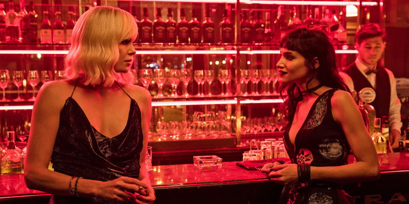 Charlize Theron and Sofia Boutella lean against a bar in red lighting. 