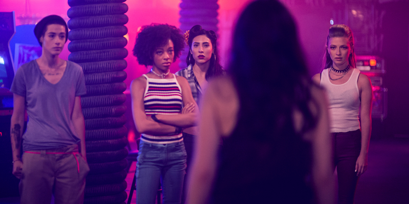 A group of lesbians drenched in pink and purple lighting look at another woman standing in front of the camera.