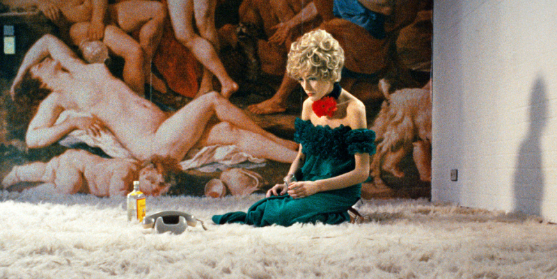 A woman in a green dress sits on her white carpet next to her phone and a bottle of alcohol. 