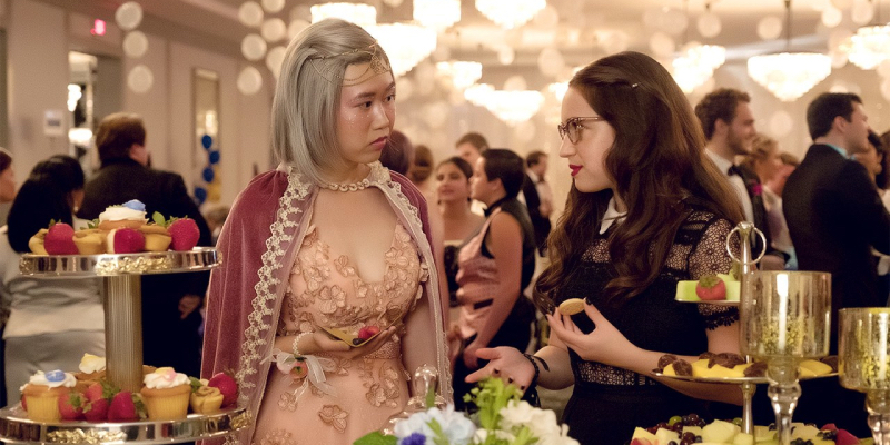 A still from Blockers. A girl in black stands by a snack table with another girl in flowery princess garb.
