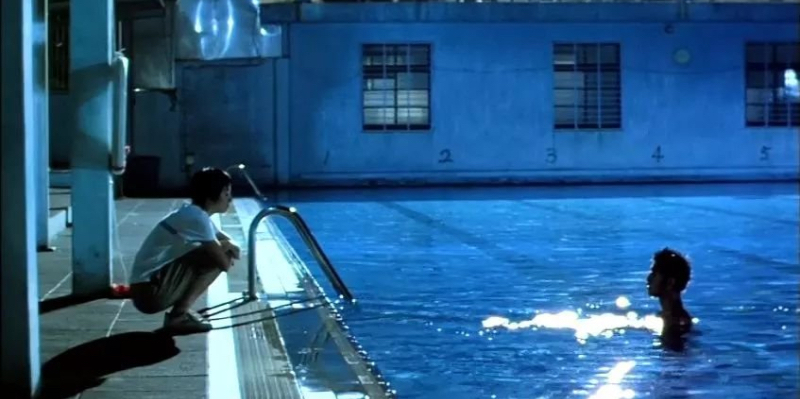 A girl crouches by a pool while talking to a boy in the pool.