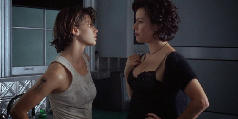 A still from the 7th best lesbian movie of all time Bound. A woman in a dirty white tank looks at a high femme in black. 