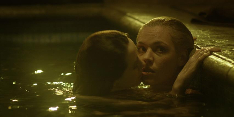 A woman kisses another woman in a pool as the other woman looks off.