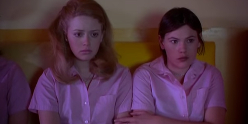 A still from the best lesbian movie of all time But I'm a Cheerleader.  Two girls watching a movie in pink outfits.