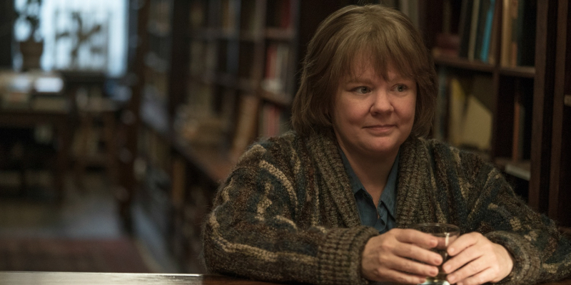 Melissa McCarthy stands in a bookstore holding a glass.