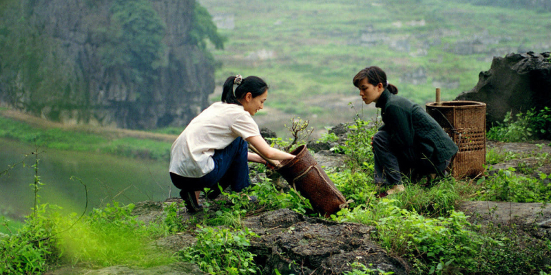 Two women crouch down in a lush green landscape.