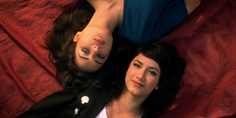 A still from Circumstance. Two young women lie on a red bed looking up toward the camera.