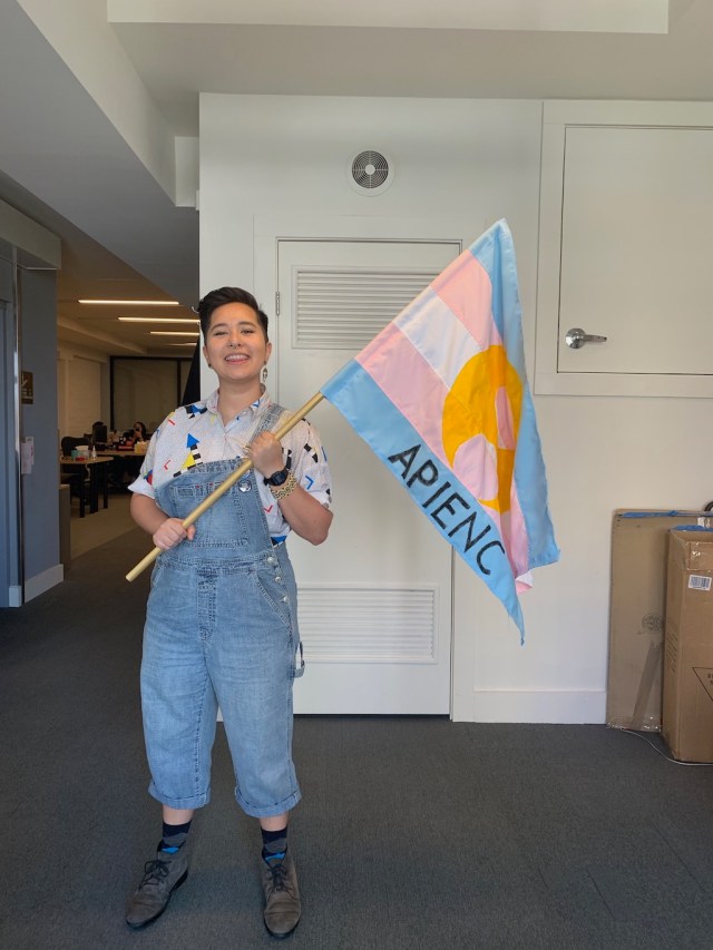 Sammie wears overalls and boots and holds up a trans APIENC flag
