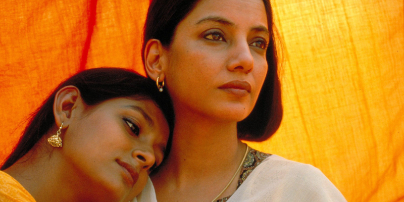 A woman rests her head against another woman with orange fabric behind them. 