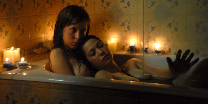Two women lie in a candle lit bathtub together.