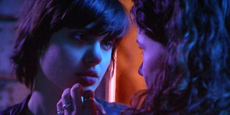 A young Angelina Jolie in bluish/purple lighting looks at another girl who reaches toward her lips.