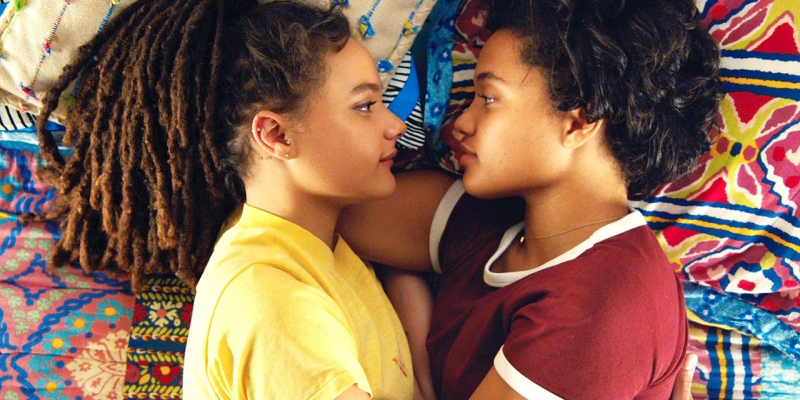Kiersey Clemons and Sasha Lane lie in bed and look at each other.
