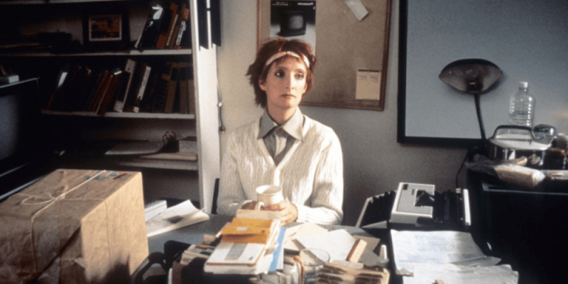 A woman in a white sweater and button down sits at a cluttered desk.