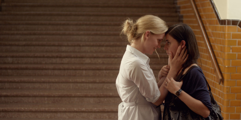 Two women almost kiss at the bottom of a staircase. 