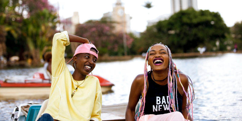 A still from the 30th best lesbian movie of all time Rafiki. Two girls, one with pink hair and the other in a pink hat, laugh while next to each other on a boat.
