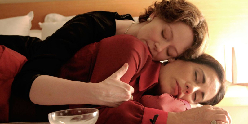 Two women cuddle on a bed with a champagne glass in the foreground.